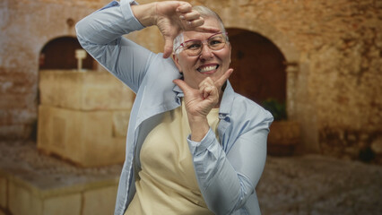 Fototapeta premium Woman with short grey hair and glasses frames face with hands on street in old town, smiling broadly in a light shirt; joyful confidence.