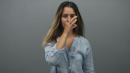 Naklejka premium Woman pinching nose and touching cheek with hand showing yellow nails in gray studio wearing loose blue shirt and long hair; disgust dismissal.
