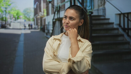 Fototapeta premium Woman with hand on chin standing on city street near stone steps and metal railings, wearing beige overshirt and white t shirt, hair tied back in ponytail, pensive pose; contemplation.