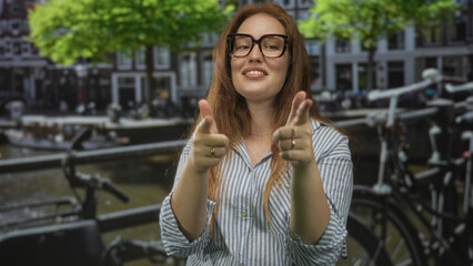 Fototapeta premium Woman wearing glasses and striped shirt points fingers to camera on street beside canal boats; playfulness.