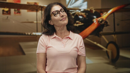 Middle age woman with glasses tilts head and looks up at vintage airplane propeller in museum building  quiet curiosity. © Krakenimages.com