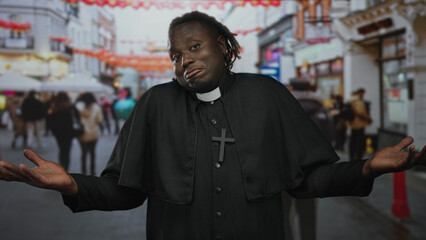 Obraz premium Man priest in black cassock and clerical collar with cross necklace shrugging with palms up on a busy street; bemusement humility.