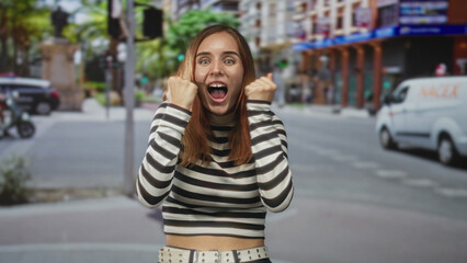 Fototapeta premium Redhead woman smiling, hands raised and showing teeth, wearing a striped top at a busy street crosswalk by buildings and cars; excitement energy.