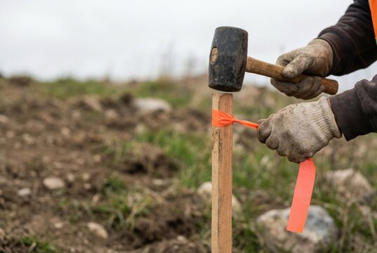 A surveyor in gloves hammers a wooden stake with an orange flagging tape into the ground.