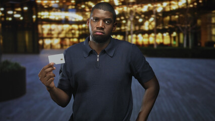 Obraz premium Man holding creditcard in right hand presenting card at lit building entrance with neutral skeptical expression and extended arm; uncertainty finance decision.
