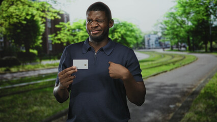 Fototapeta premium Man holding a credit card in left hand and points finger to card near his chest on a tree lined urban street with tram tracks; doubt identity security.