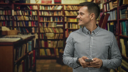 Man with moustache holding smartphone in hands and looking to the side in a building filled with bookshelves and rows of books  quiet curiosity. © Krakenimages.com