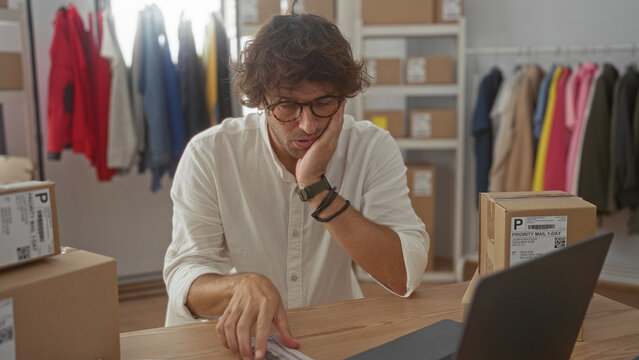 Man counting cash and reviewing packages at a warehouse packing station, later clutching his head in hands and staring at laptop; financial stress.
