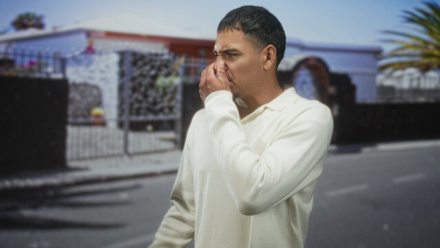 Man pinches nose in white sweater on a residential street while reacting to a strong odor; disgust odor.