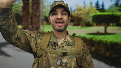 Man in camouflage uniform and dog tag necklace waving hand on street with palm trees; duty and...