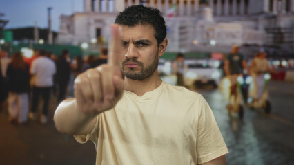 Fototapeta premium Young hispanic man pointing index finger directly at camera in a crowded urban street at dusk; serious.