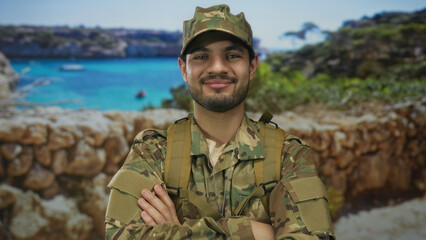 Man in camouflage military uniform stands with arms crossed on street by coastal rocks under bright...