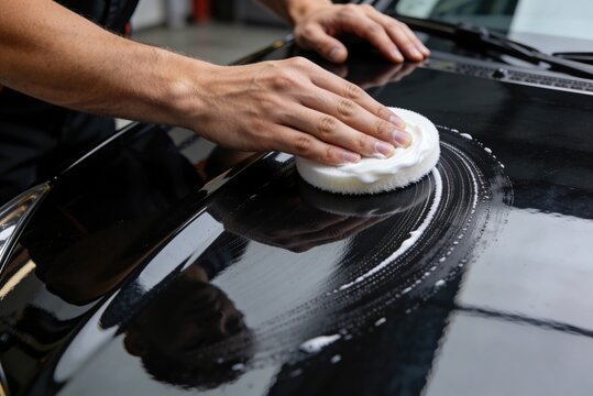 A detailer's hand polishing the black hood of a car with a white foam applicator pad and wax.