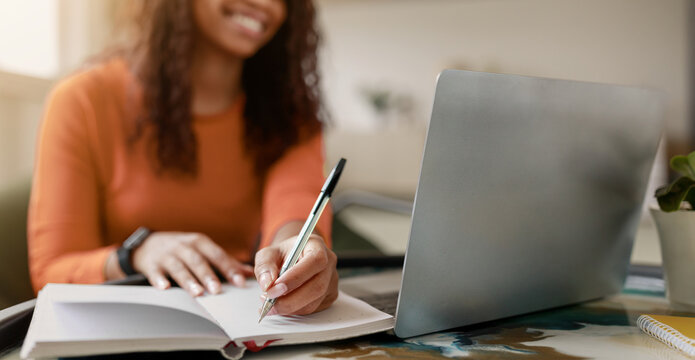 A woman sits at a desk with a laptop open in front of her. She writes in a notebook with a pen, focusing on her task. The sunlight enters the room, creating a warm environment.