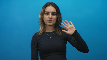 Fototapeta premium Woman raising her hand and waving in studio against a bright blue backdrop wearing a black top; friendly greeting.