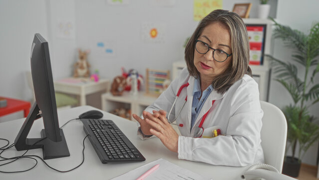 Woman doctor with stethoscope and hands clasped at computer desk in pediatric clinic; calm compassion.