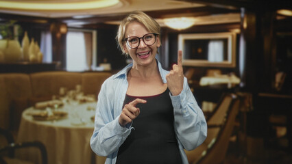 Fototapeta premium Blond woman smiling with glasses, eyes closed, crossing fingers and pointing with hands while wearing casual shirt in restaurant; hopeful.