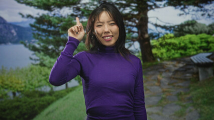 Woman points finger upward beside her face on a forest lakeside path, wearing purple turtleneck and hoop earring  serenity. © Krakenimages.com
