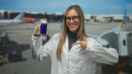 Fototapeta premium Woman smiling, holding blue bottle and points finger to bottle at airport gate beside airplane jetbridge; happiness wellness health.