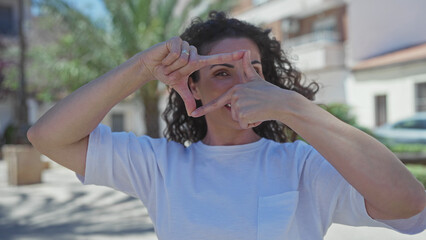 Young hispanic woman wearing white shirt framing view with hands on street under bright daylight; creative vision.