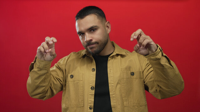 Man wearing tan jacket holds air quotes gesture with both hands in studio against red background; skepticism.
