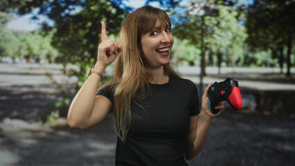 Woman wearing black t shirt holding red and black game controller points index finger up on a forest path smiling broadly; playful gaming moment.