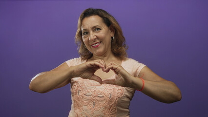 Obraz premium Hispanic middle aged woman with soft smile making heart shape gesture with hands in purple studio; love.