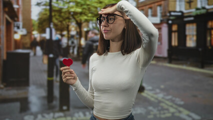 Young woman wearing glasses and a white long sleeve top holds a small red heart and shields her eyes with hand on forehead on a street lined with shops  longing romance. © Krakenimages.com
