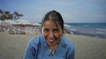 Young hispanic woman in blue attire laughing and enjoying a sunny day at the beach, with palm trees and ocean waves creating a vibrant seaside background scene.