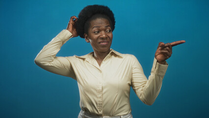 Woman scratches head and points finger in studio against blue backdrop wearing beige shirt with...