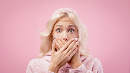A woman stands with wide eyes and covers her mouth with her hands. She appears surprised or shocked. The background is pink, and she wears a cozy outfit. © Prostock-studio