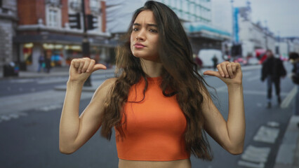 Fototapeta premium Hispanic teenage woman points both thumbs toward herself on a busy street lined with shops and moving vehicles; self doubt.