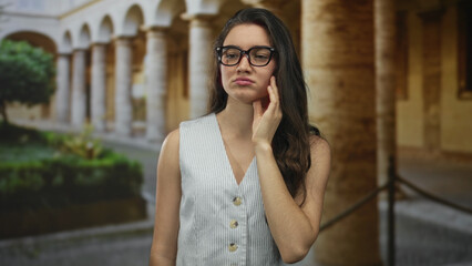 Fototapeta premium Young hispanic teenage woman stands with hand on cheek for toothache in old town street by ancient building; discomfort.