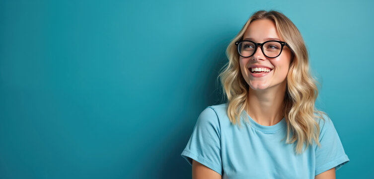 Young woman with blond hair wears glasses and blue t-shirt. She smiles brightly against a solid blue studio background. This portrait conveys positivity, confidence and intelligence.
