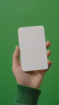 A video mockup of a hand holding a blank white vertical card against a green background. This simple footage is ideal for showcasing business cards, flashcards, or small flyers.