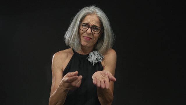 Woman with outstretched hands making pleading gesture in studio against a black wall; uncertainty asking help.