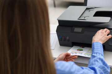 Woman taking printed sheet of paper from printer at white desk indoors, closeup