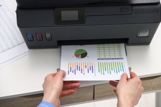 Woman taking printed sheet of paper from printer at white table, above view