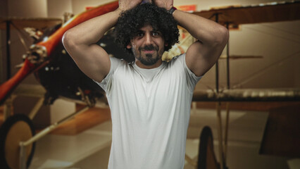 Man with curly hair and beard, hands on head beside vintage biplane in museum exhibit, wearing white tshirt and showing biceps  nostalgia amusement. © Krakenimages.com