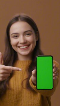 A vertical video mockup of a happy woman pointing at a smartphone with a green screen. This realistic footage is ideal for user testimonials, app promotions, or sales.