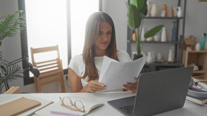 Woman holds and reads document, hand gesture over laptop at studio desk with glasses, notebook and plant shelves; work concentration.