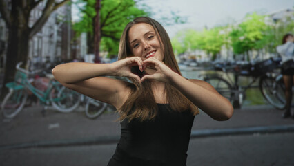 Fototapeta premium Woman forming heart with hands and smiling on street by canal with parked bicycles and trees; love warmth.