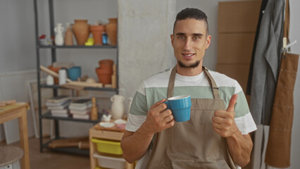 Fototapeta premium Man in apron holding a blue mug with both hands and showing a thumbs up, looking thoughtful in a pottery studio with shelves of clay pots; quiet pride.