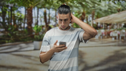 Fototapeta premium Man holding smartphone with both hands, tapping and scrolling the screen, wearing striped tshirt and beard, standing on street cafe walkway under palm trees; contemplation.