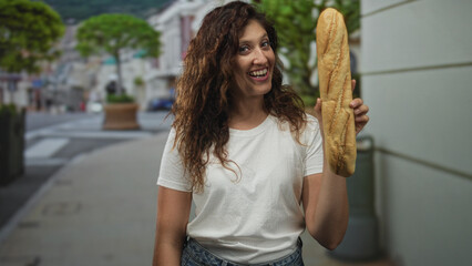 Woman holds baguette upright on street while smiling and showing hands in a city sidewalk scene  everyday joy. © Krakenimages.com