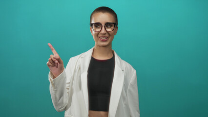 Naklejka premium Woman with glasses and short buzzcut wearing white jacket and black crop top raises fists and smiles in studio; victory joy.