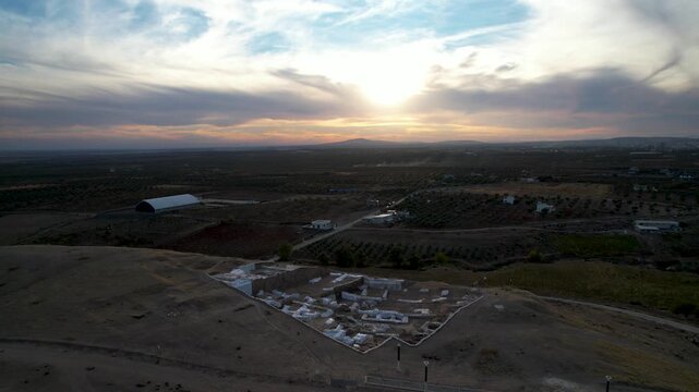 Aerial drone shot of the large archaeological mound of Oylum Hoyuk at sunset, under dramatic clouds, featuring the modern white protective roof over the Aidesim Mosaic Basilica site.
