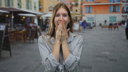 Fototapeta premium Young blonde woman cups cheeks with hands while smiling at a street restaurant terrace outdoors; happiness.