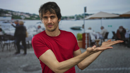 Fototapeta premium Man with open palms gesture wearing red tshirt on waterfront cafe terrace overlooking boats and bay; invitation confusion.