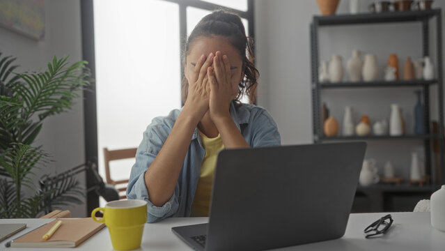 Woman covers face with hands at laptop in studio, seated at a cluttered desk with yellow mug, notebook, glasses and potted plant; stress fatigue.